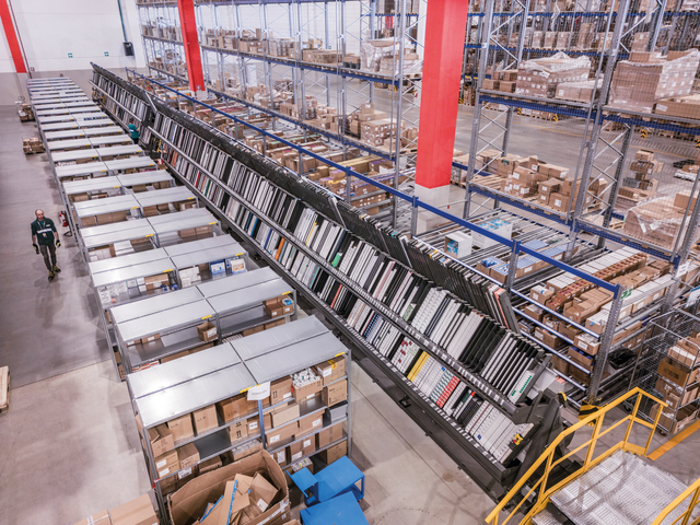 A large warehouse with shelves full of boxes, a conveyor system, and a worker walking along the aisle for pharmaceutical distribution.