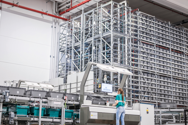 A woman operates a control panel in a modern, automated warehouse for pharmaceutical distribution with tall shelves and sorting bins.