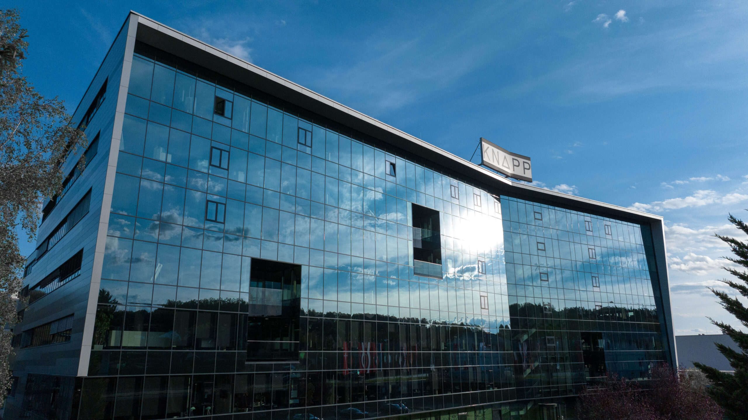 Modernes gläsernes Bürogebäude mit blau getönten Fenstern, in denen sich der Himmel spiegelt; auf dem Dach befindet sich ein Schild mit der Aufschrift APP News".