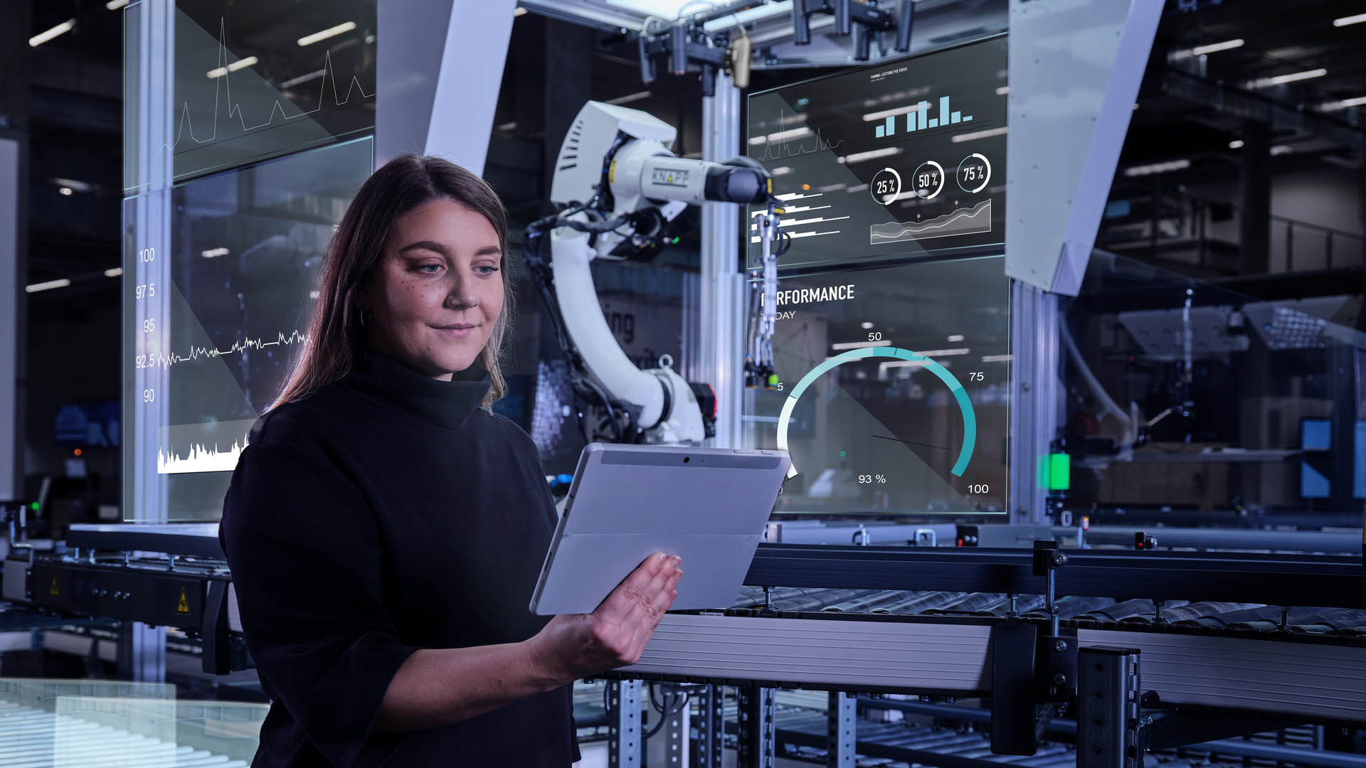 Frau mit Tablet in einer Hightech-Fabrik bei KNAPP auf der LogiMAT, umgeben von Datenanzeigen und einem Roboterarm im Hintergrund.