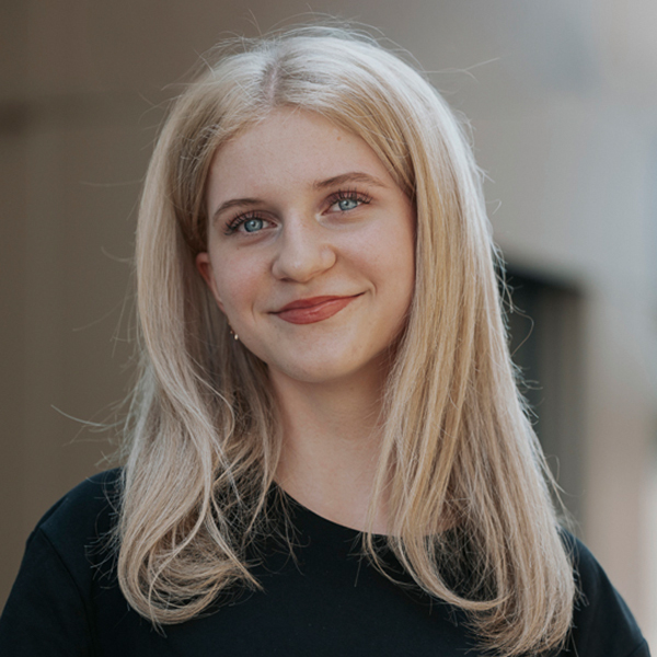 Eine junge Frau mit langem, glattem blondem Haar lächelt sanft. Sie trägt ein schwarzes Oberteil.