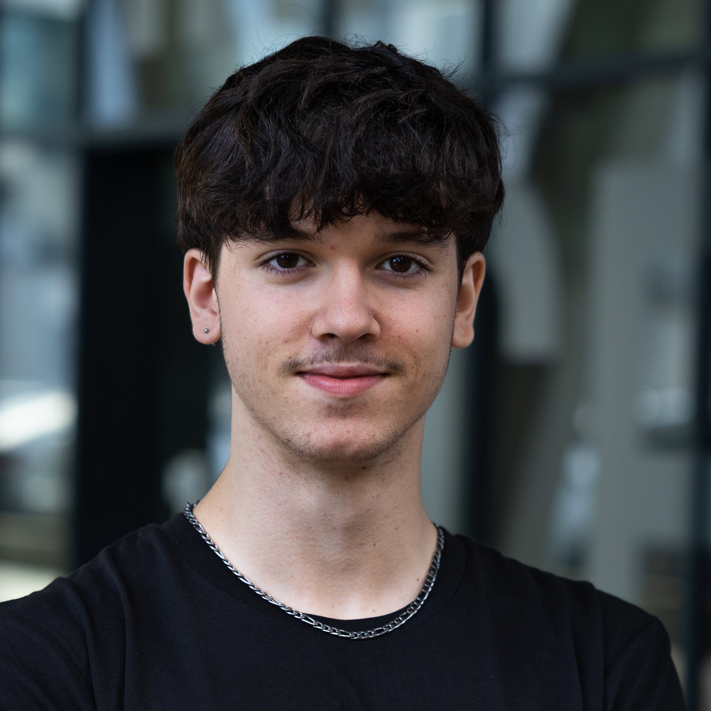 Ein junger Mann mit kurzem, dunklem, lockigem Haar und einem leichten Lächeln steht im Freien, trägt ein schwarzes T-Shirt und eine silberne Halskette.
