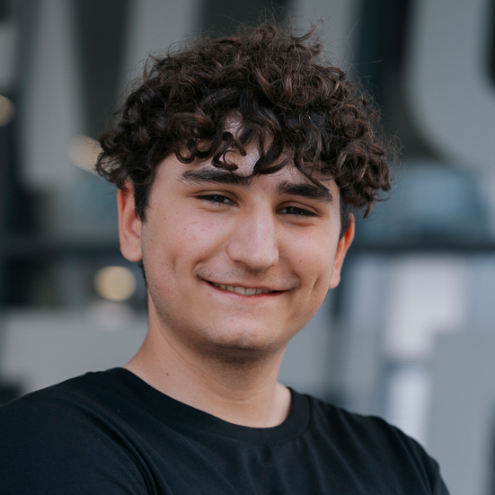 Ein junger Mann mit lockigen braunen Haaren und einem schwarzen T-Shirt lächelt sanft in die Kamera.