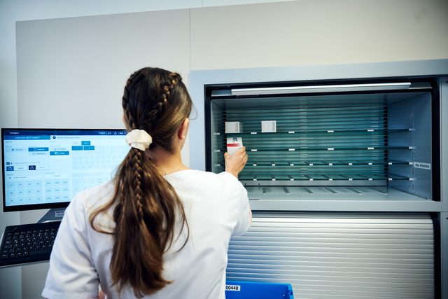 Eine Frau in einem weißen Kittel stellt einen Behälter in ein medizinisches Lager - Victorinox setzt auf zentrale Verteilung - und zeigt die Daten auf einem Computerbildschirm in der Nähe an.