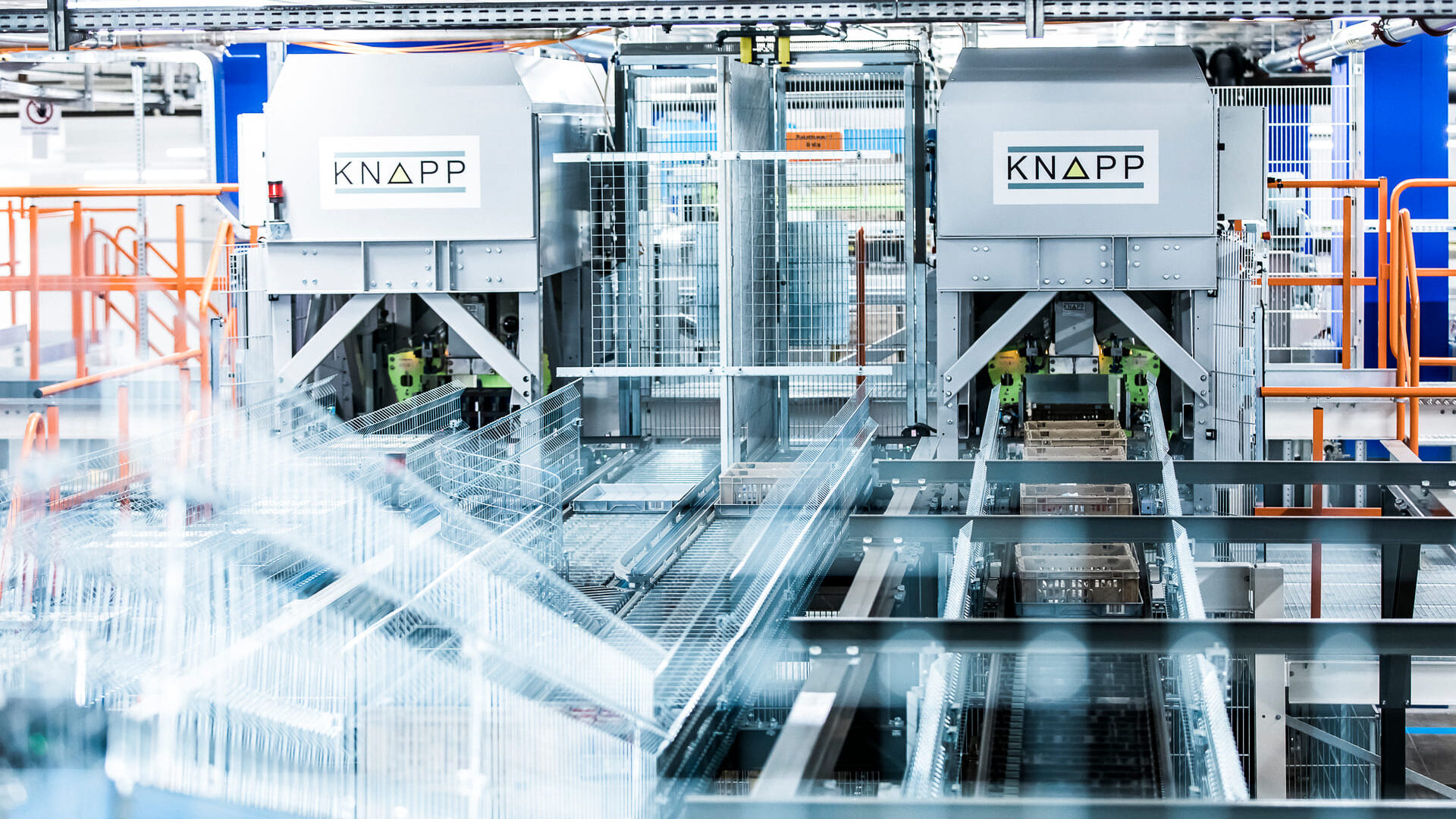KNAPP Food Solution Migros Zurich Un système moderne d'entrepôt automatisé avec des rayonnages métalliques, des bandes transporteuses et deux grandes machines étiquetées "KNAPP", avec un éclairage vif et des barrières de sécurité orange dans un environnement industriel.