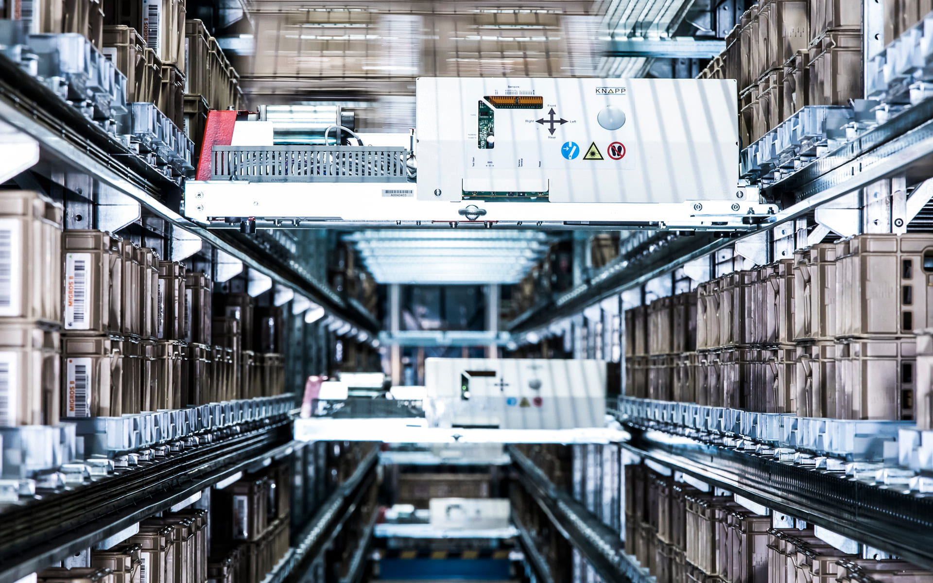 Vue rapprochée d'un système de stockage et de récupération automatisé dans un entrepôt, avec des étagères de boîtes brunes et un mécanisme robotique se déplaçant sur des rails métalliques entre les étagères.