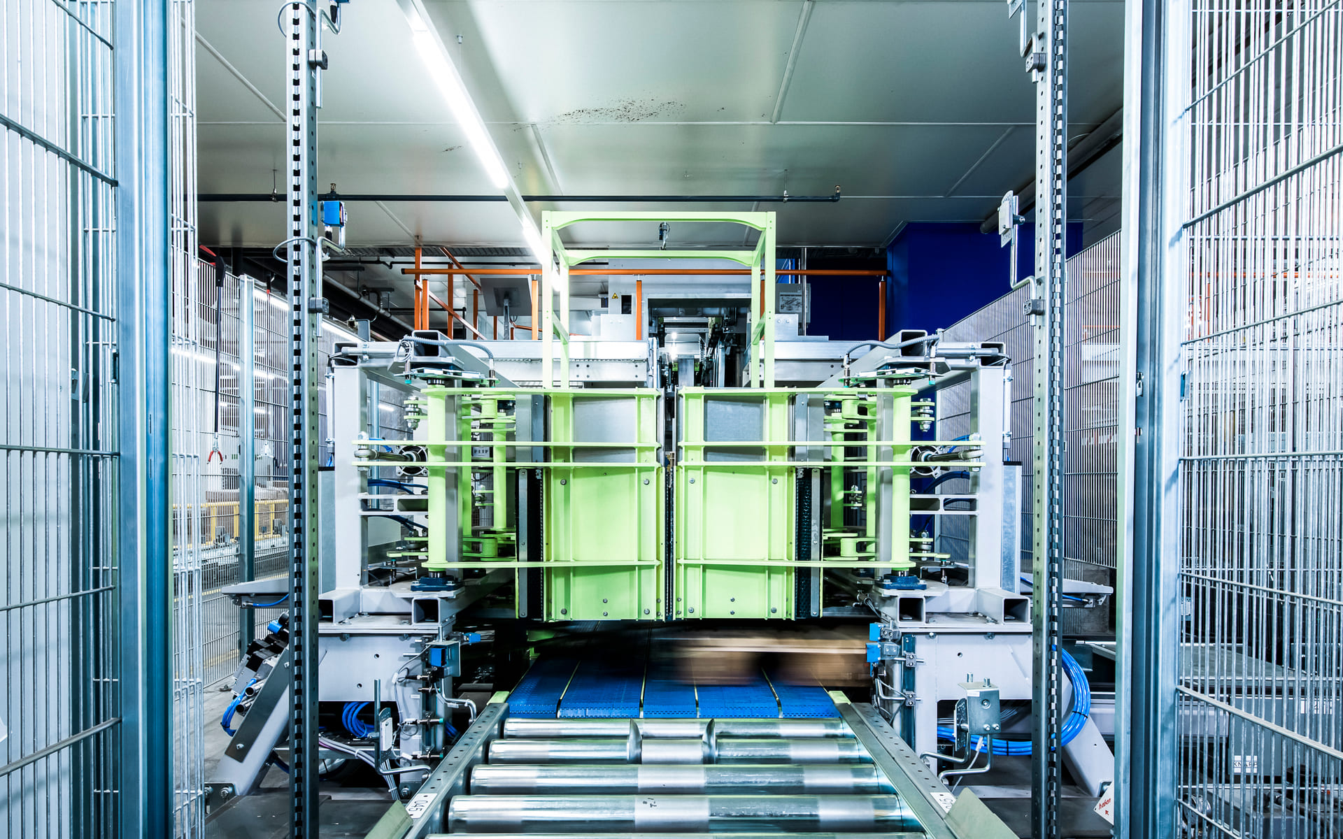 Une machine industrielle moderne avec des rouleaux métalliques et un cadre vert à l'intérieur d'une usine bien éclairée, entourée d'une clôture métallique, utilisée pour la manutention ou le traitement automatisé de matériaux.