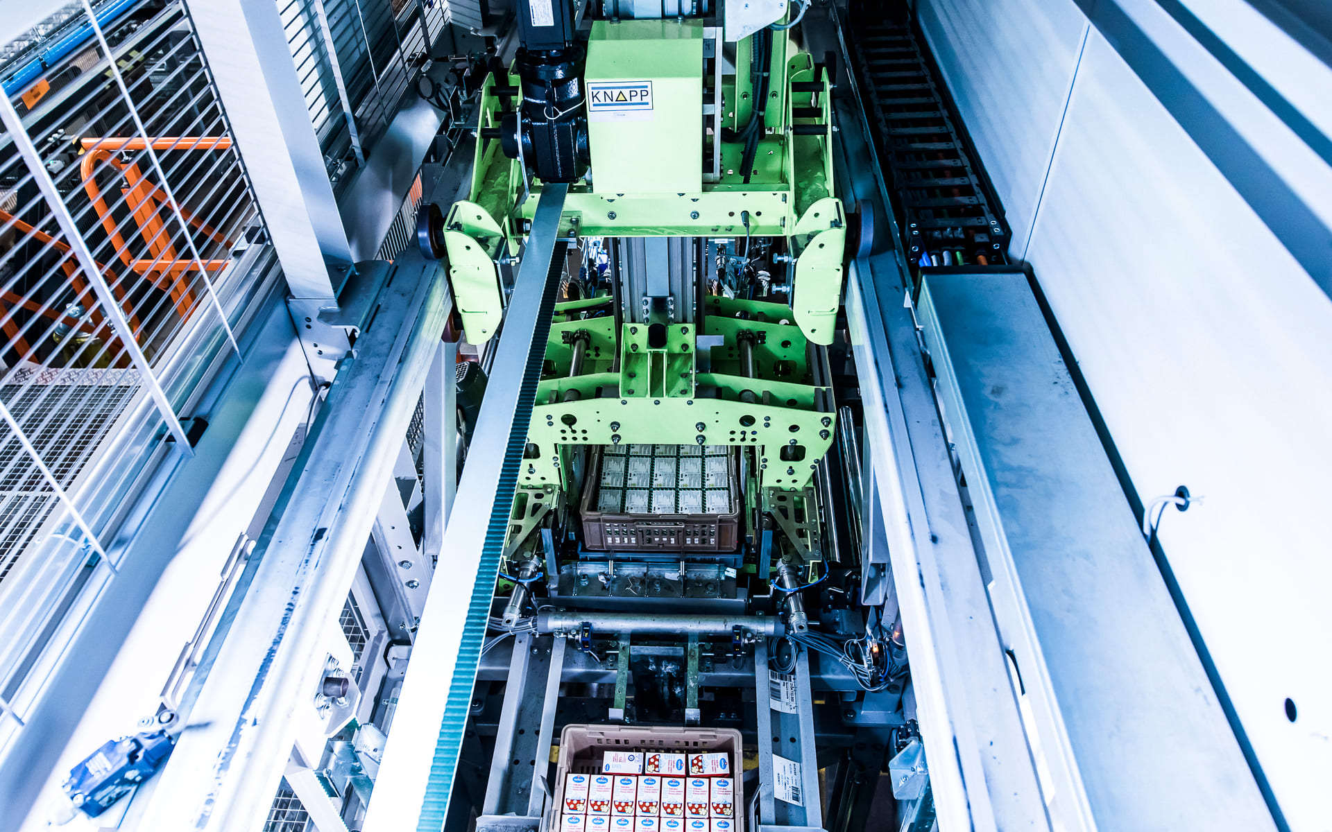 Vue aérienne d'une machine industrielle automatisée manipulant des paquets dans un entrepôt, avec des rangées de produits emballés sur un système de convoyeurs et une structure métallique de chaque côté.