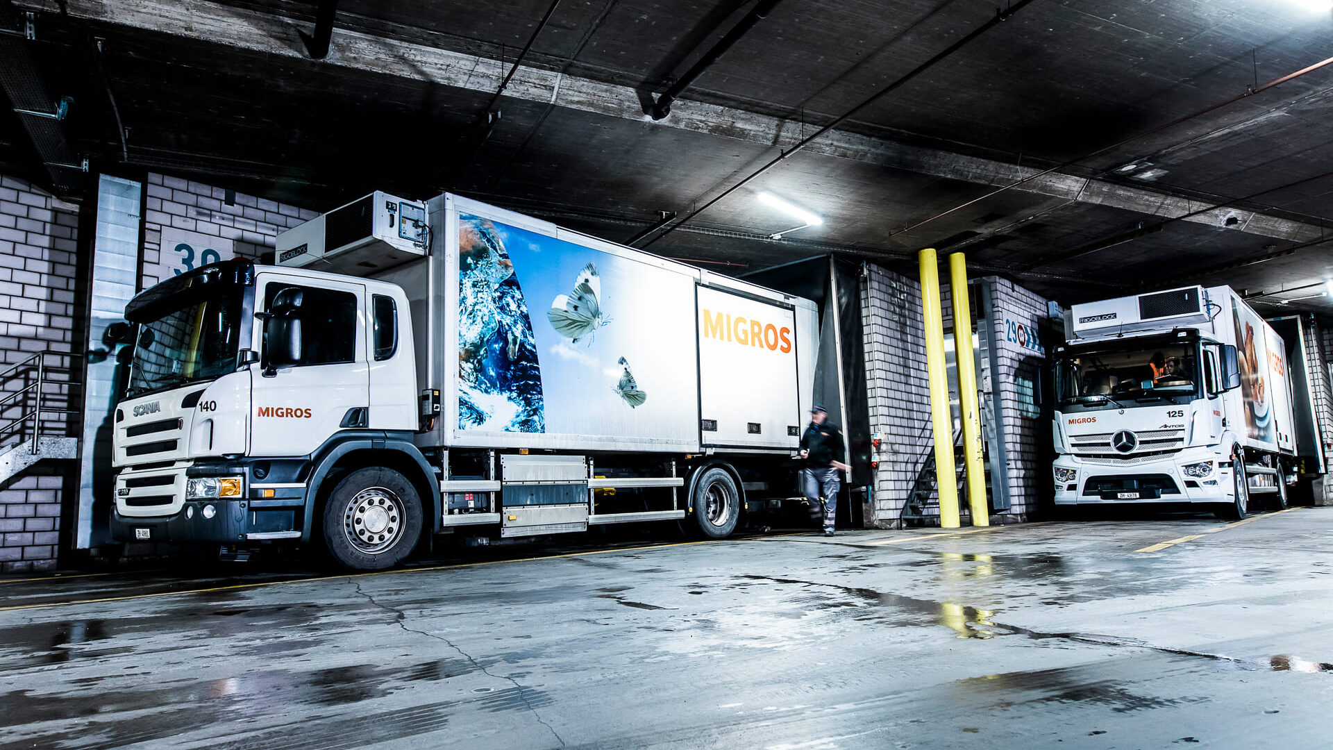 KNAPP Food Solution Migros Zürich Deux camions de livraison Migros sont garés à l'intérieur d'un quai de chargement, se reflétant sur le sol mouillé sous des lumières vives. Une personne se tient à côté d'un camion dans cette scène industrielle moderne où Migros Zurich automatise la livraison de 150 magasins.