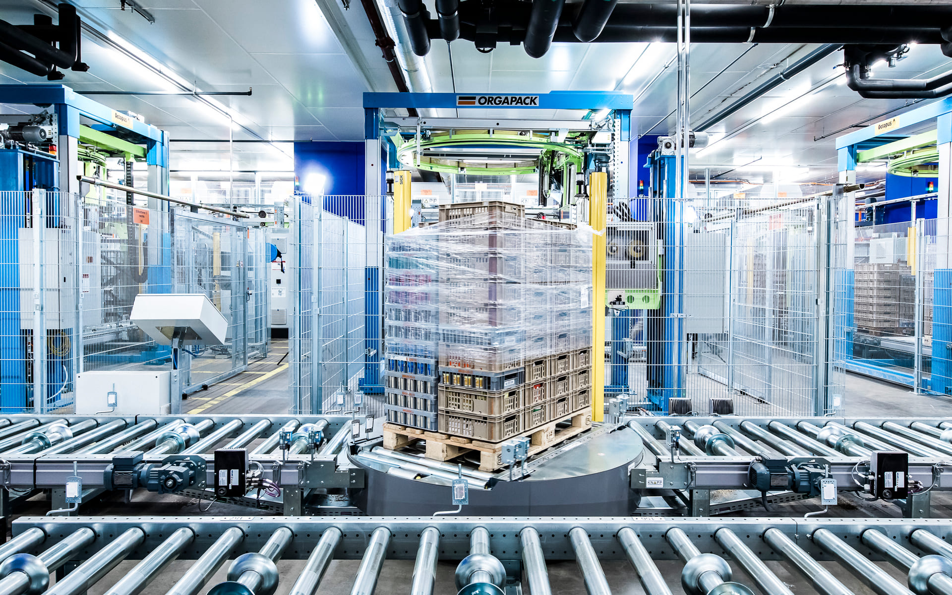 Une palette empilée de marchandises est emballée dans un film plastique par des machines automatisées dans une usine moderne et lumineuse, dotée de rouleaux métalliques et d'une clôture bleue.
