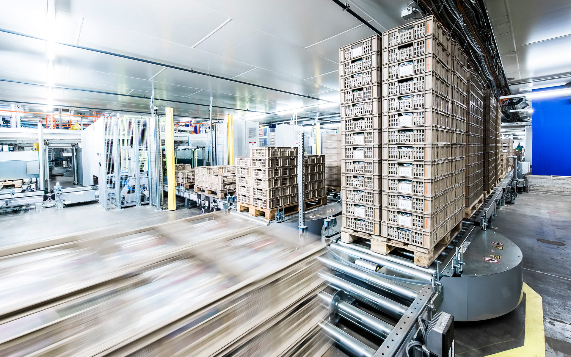 Un entrepôt moderne automatisé avec des piles de caisses sur des palettes se déplaçant sur des tapis roulants, entouré de machines industrielles et d'un éclairage intense.