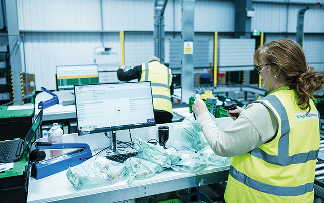 Person arbeitet an einer Logistikarbeitsstation mit Monitor, Verpackungsmaterial und Fördertechnik in einer automatisierten Lagerumgebung.