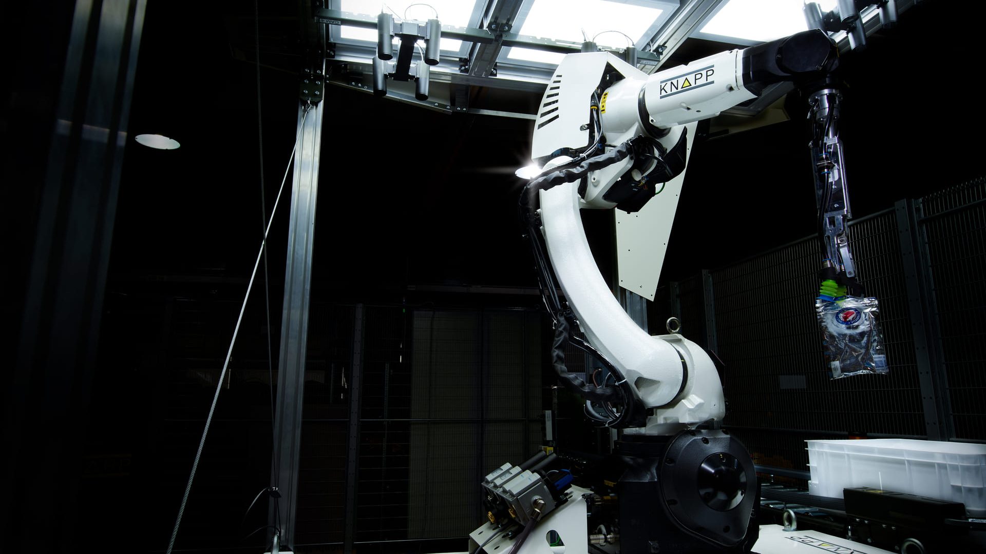 Un brazo robótico etiquetado como "KNAPP" opera en un oscuro entorno industrial, recogiendo una pequeña bolsa de plástico que contiene artículos de colores bajo brillantes luces cenitales.