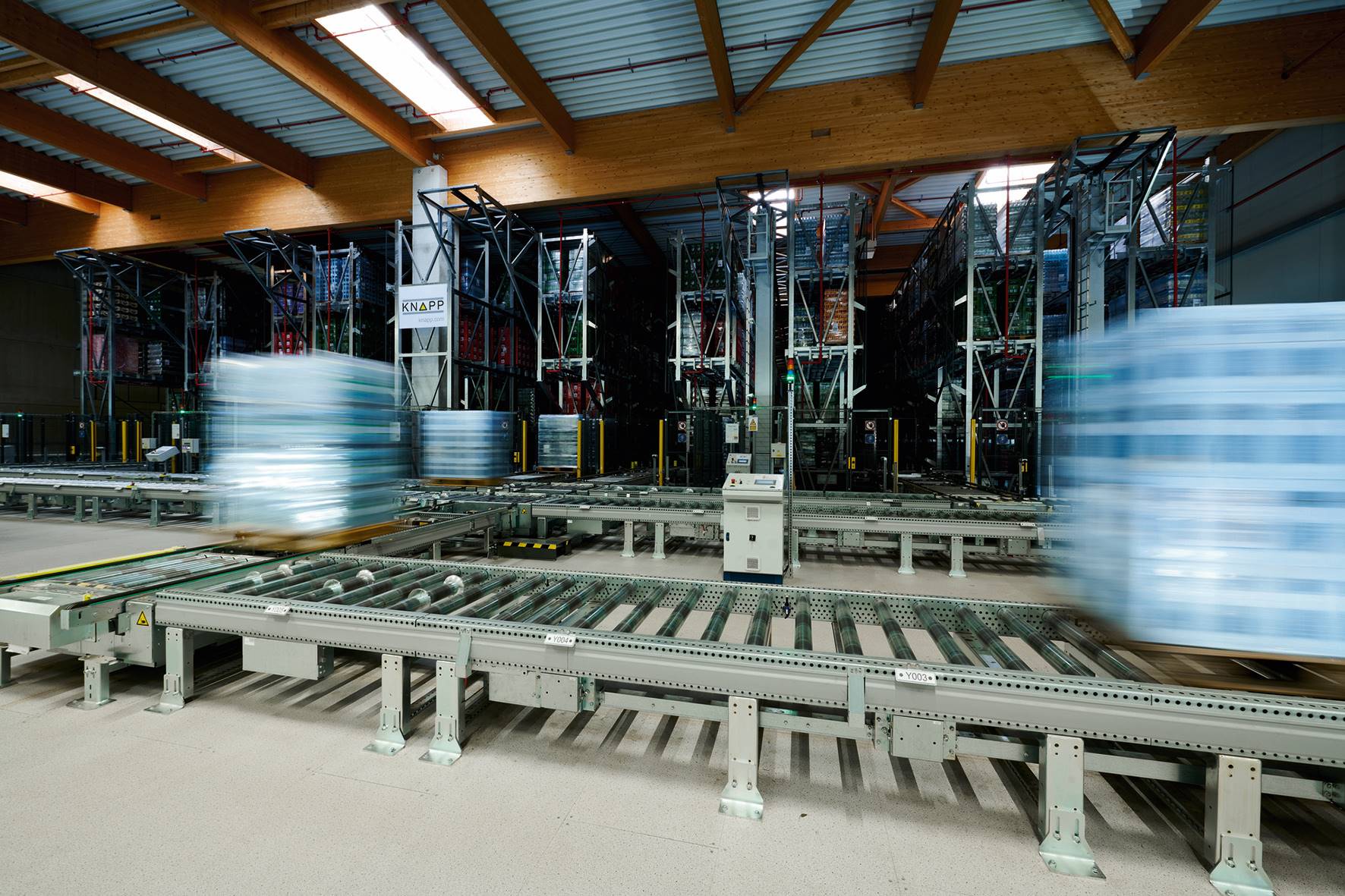 Automatisiertes Lager mit Förderbändern und Hochregalen, in Plastik verpackte Paletten, die sich schnell durch das System bewegen und einen unscharfen Bewegungseffekt unter einer hohen Decke mit Holzbalken erzeugen.