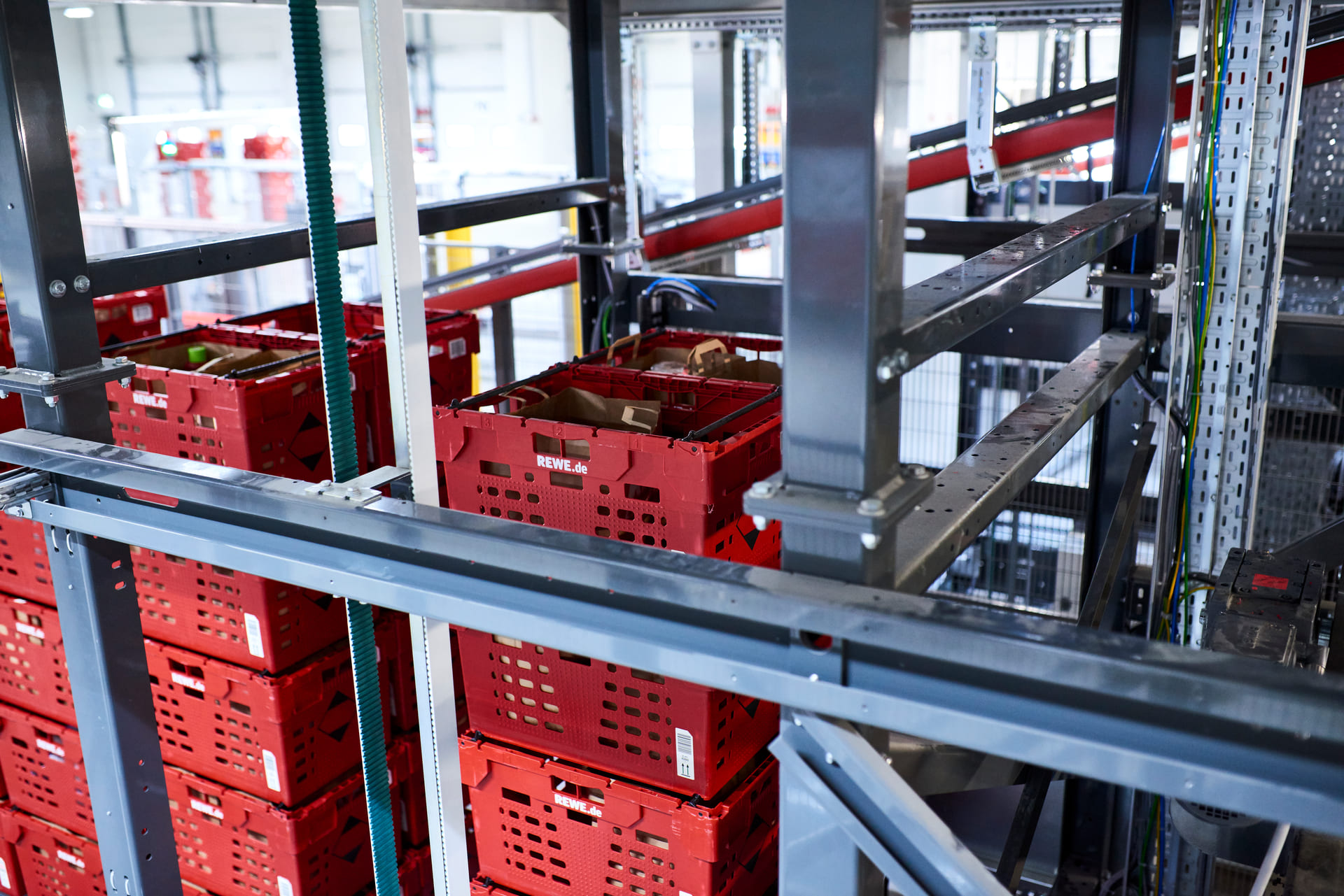 Des caisses en plastique rouge sont empilées et organisées sur des étagères métalliques dans un entrepôt, avec quelques caisses contenant des marchandises et un système de convoyeur à bande visible à l'arrière-plan.