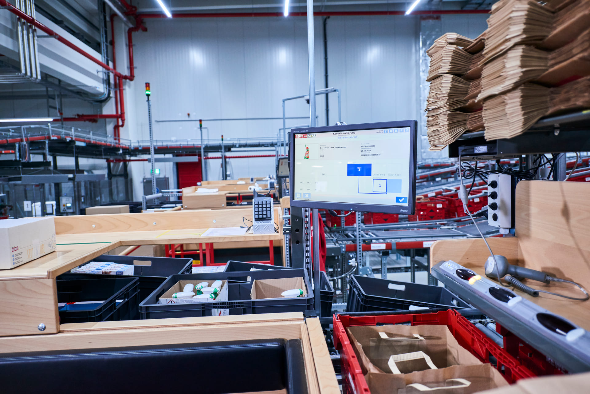 Poste d'emballage d'un entrepôt moderne avec un écran d'ordinateur affichant des instructions d'emballage, entouré de cartons, de sacs emballés, de codes-barres et de divers colis sur des surfaces en bois. Des étagères et des équipements sont visibles à l'arrière-plan.