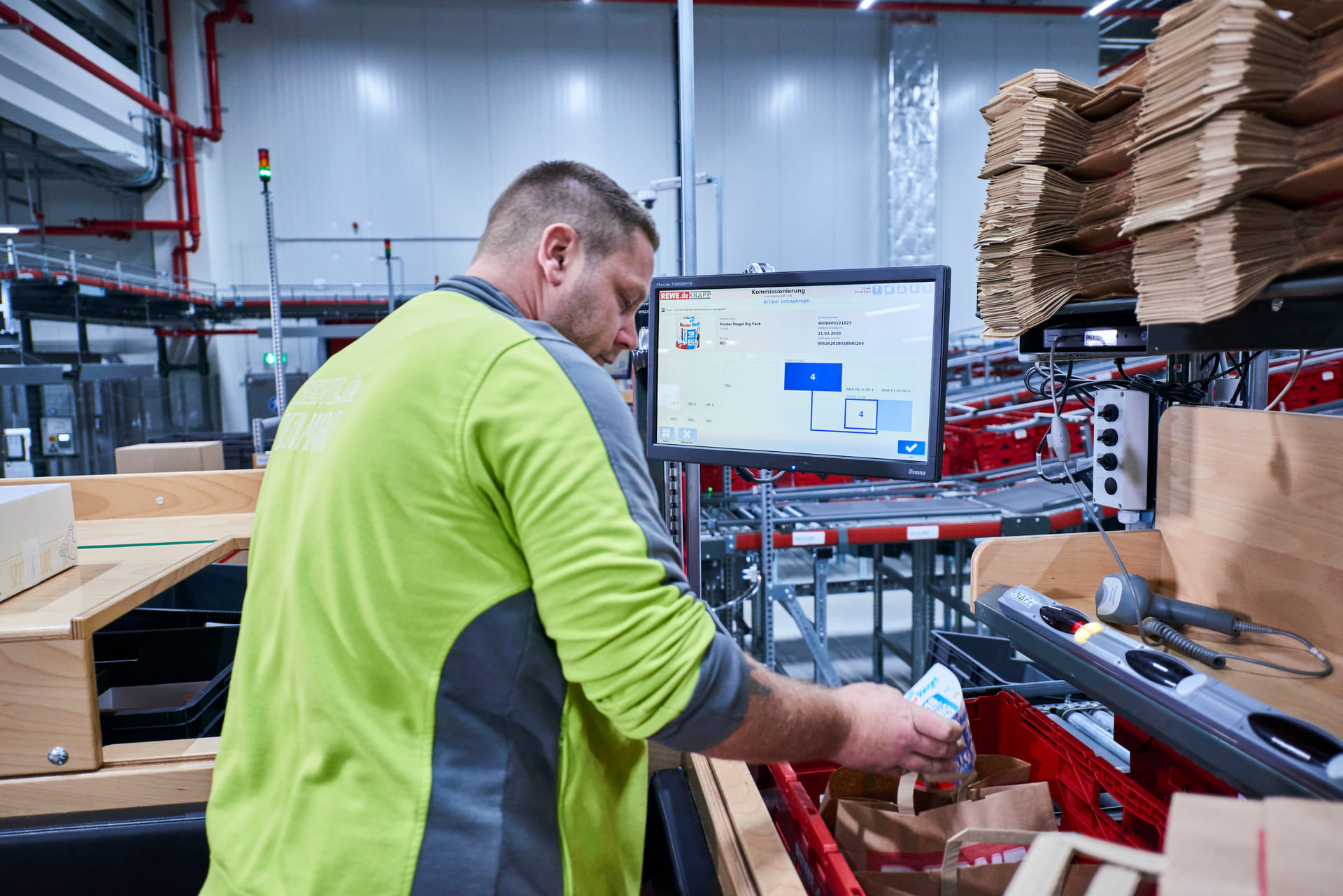 Un ouvrier en chemise verte met des articles dans un sac sur un poste de travail industriel, avec des étagères, des outils et un ordinateur affichant des informations logistiques dans un entrepôt.