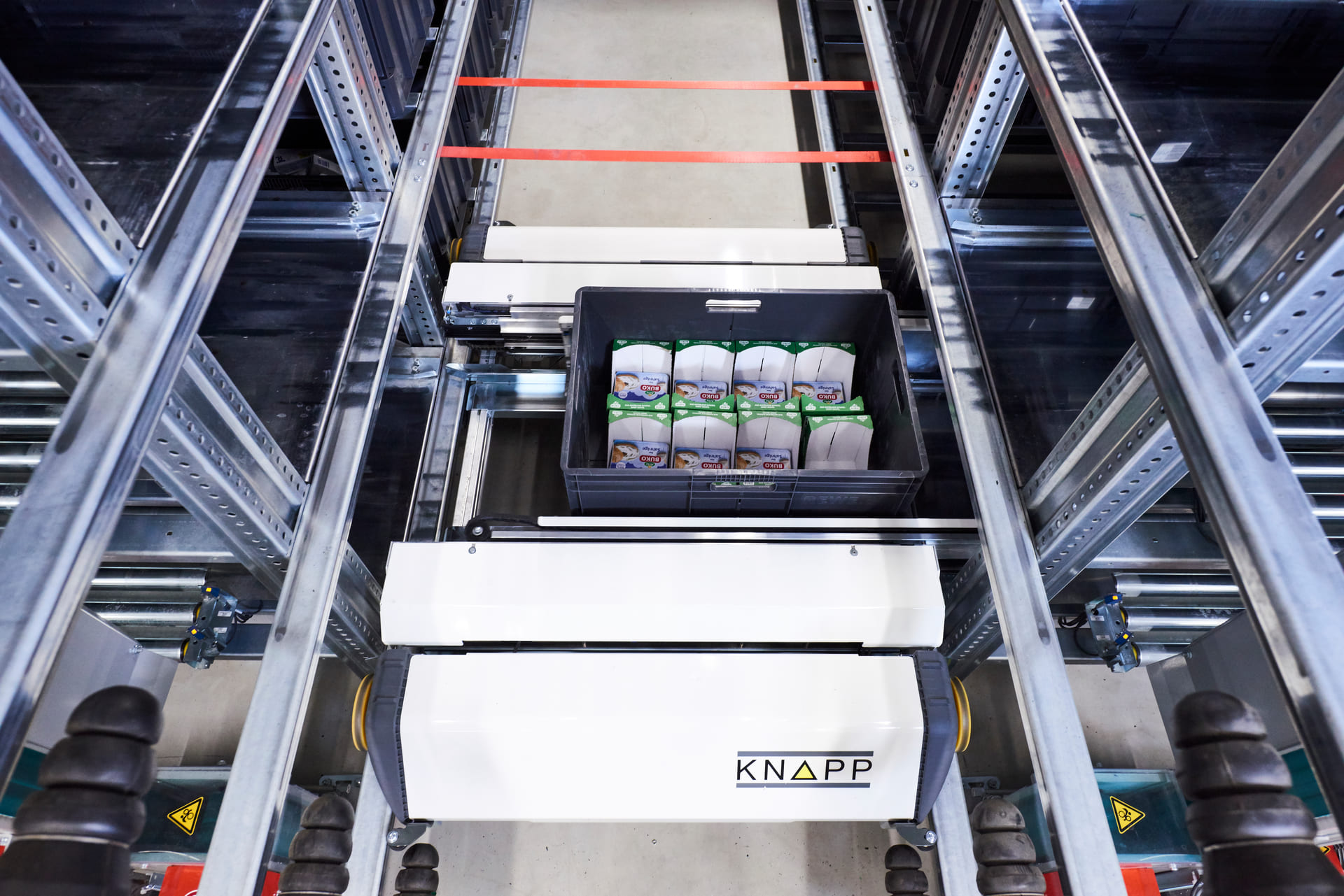 A KNAPP automated storage and retrieval system, doté de technologies de pointe pour l'e-commerce alimentaire, récupère une caisse noire remplie de produits emballés depuis les étagères métalliques d'un entrepôt.