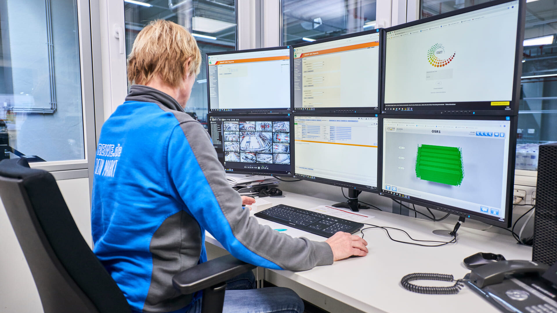 Salle de contrôle Dans une salle de contrôle moderne, une personne est assise à un bureau devant six écrans d'ordinateur affichant des tableaux, des graphiques et des images de caméras de sécurité. La personne porte une veste bleue et grise.
