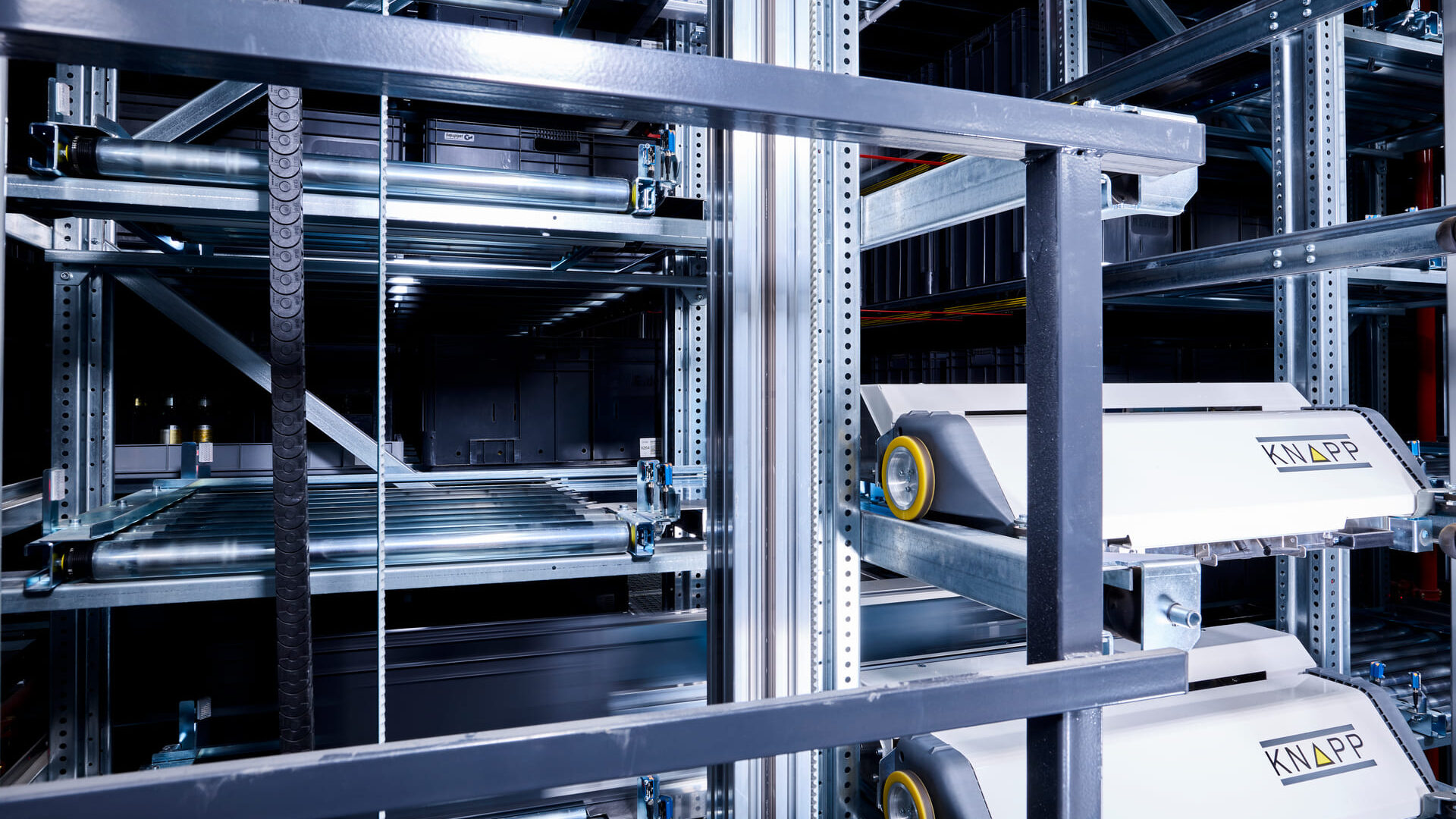 Evo Shuttle de KNAPP Système d'entrepôt automatisé composé de rayonnages métalliques, de bandes transporteuses et de deux navettes robotisées blanches étiquetées "KNAPP", utilisées pour transporter des marchandises dans la zone de stockage.