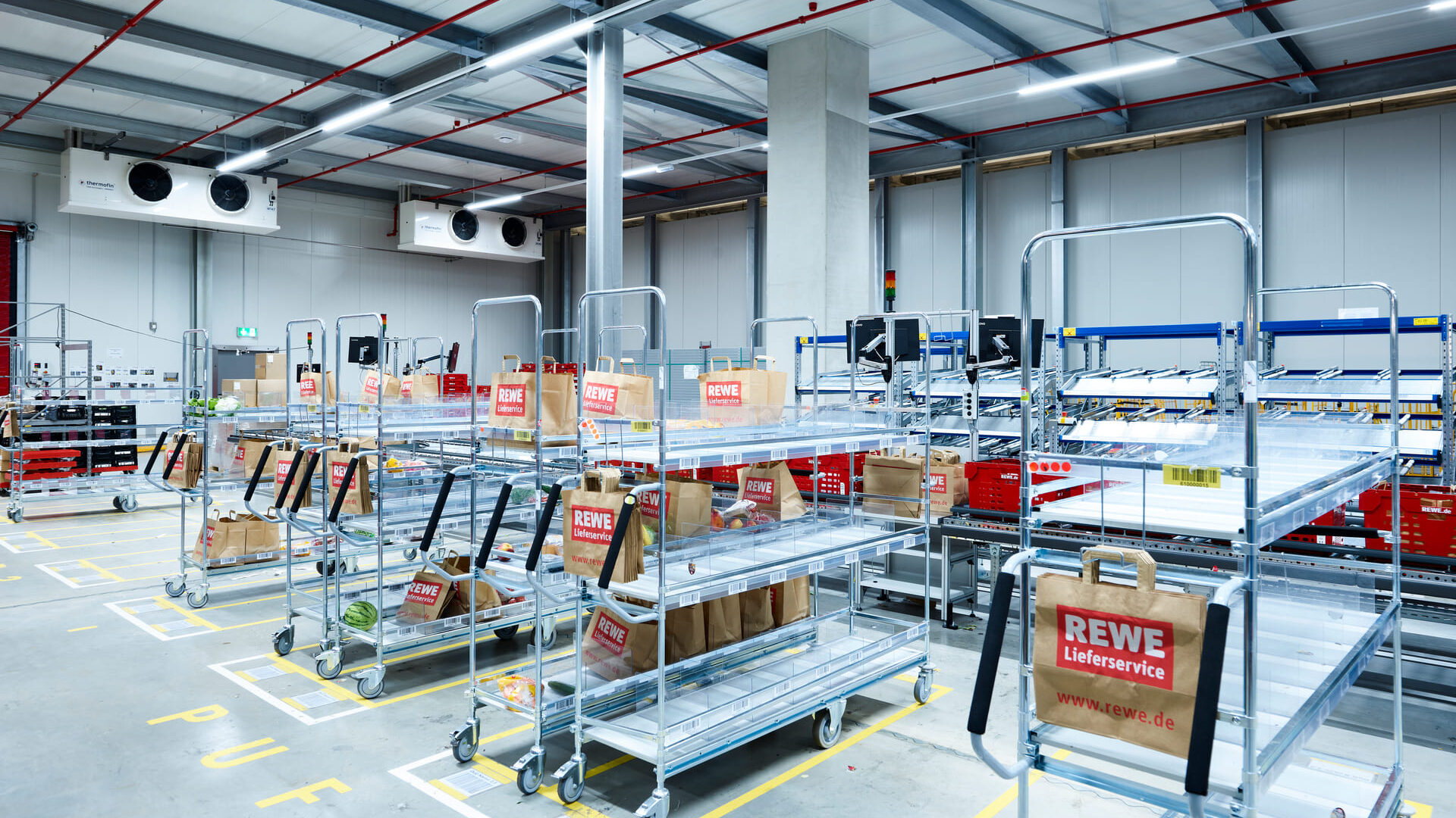 Produits alimentaires Un entrepôt avec des chariots roulants en métal contenant des sacs en papier étiquetés "REWE Lieferservice". L'espace est bien éclairé, les étagères et les caisses sont bien rangées et les chariots vides sont alignés sur des espaces délimités au sol.