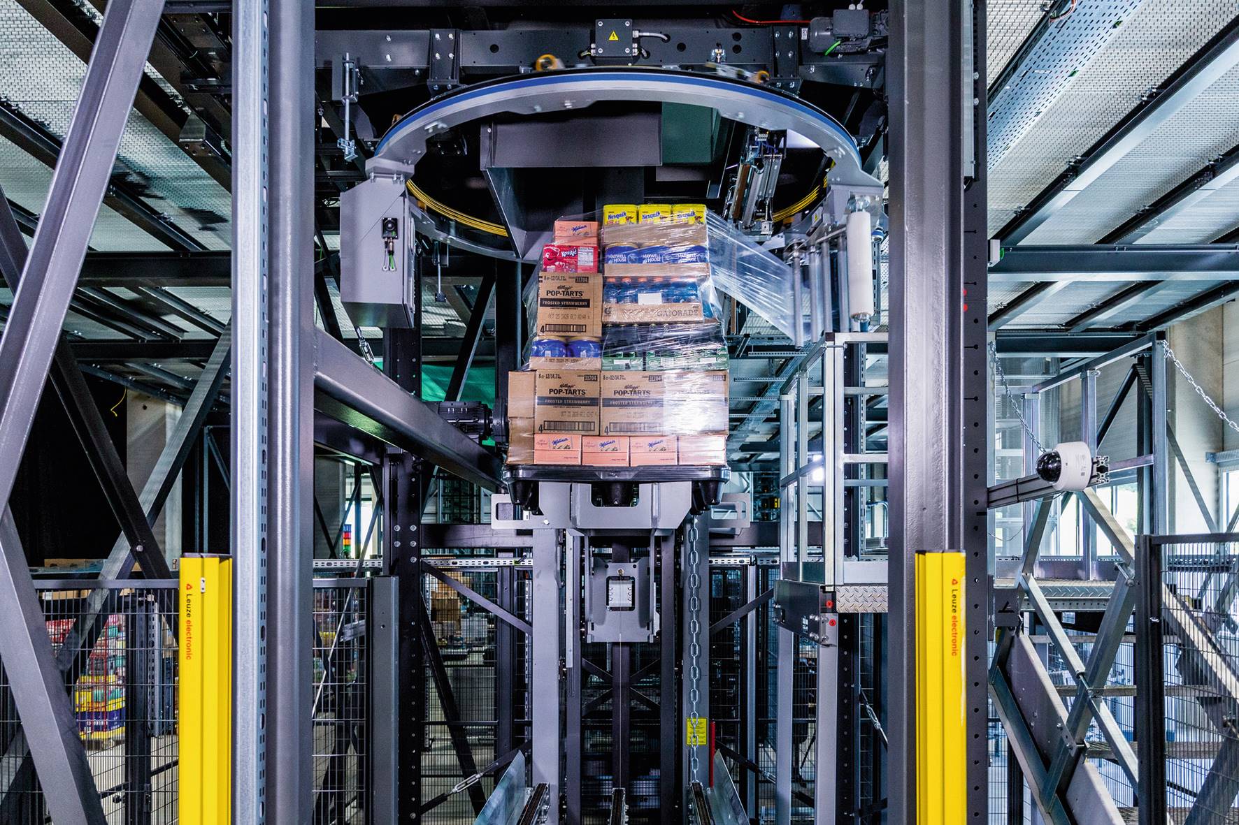 Ein automatisiertes Robotersystem hebt und transportiert gestapelte Kisten mit verschiedenen Waren auf einer Plattform innerhalb eines Lagers, die von Metallträgern und Sicherheitsbarrieren umgeben ist.