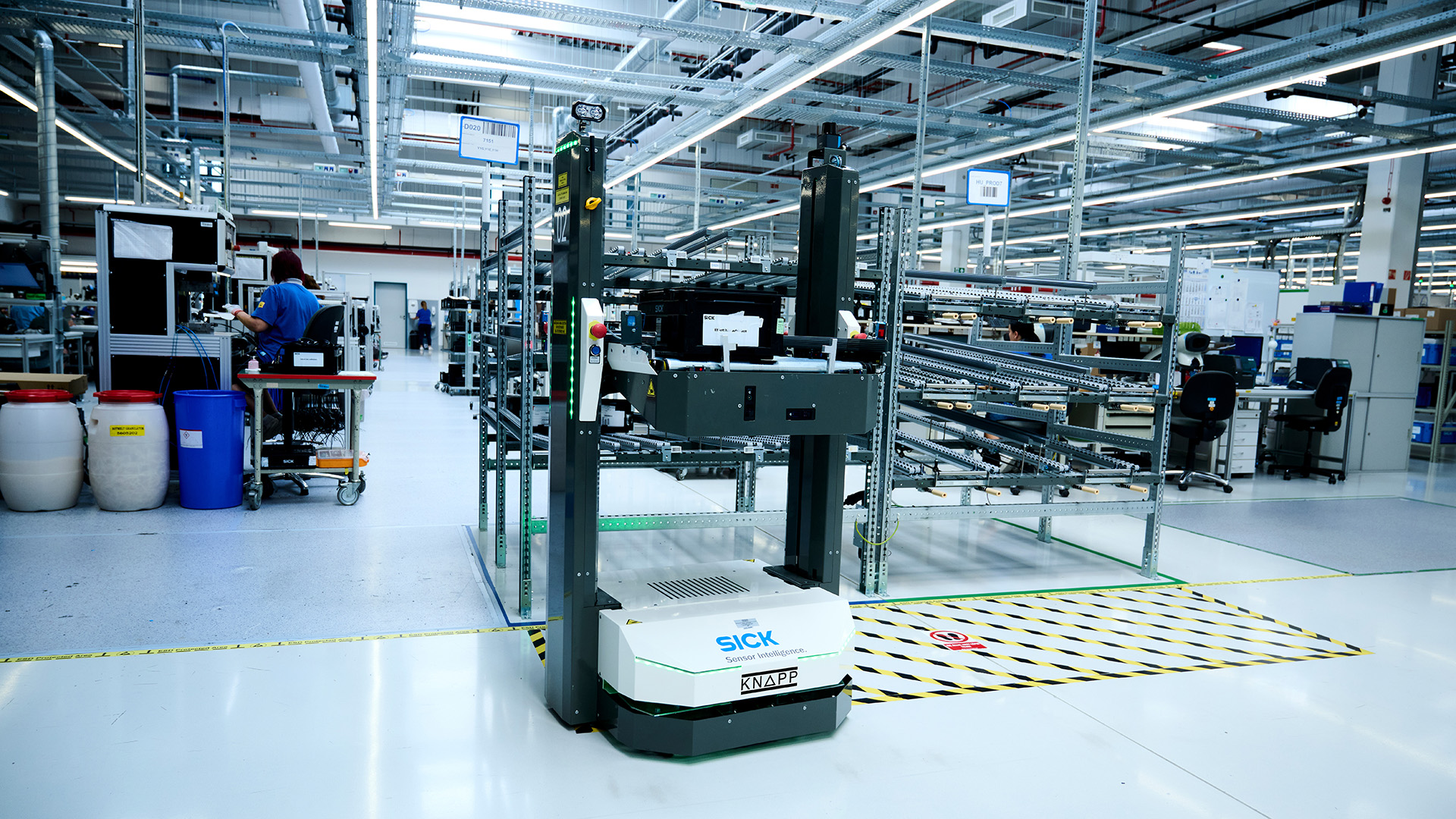 Ein autonomer mobiler Roboter mit den Logos von SICK und KNAPP bewegt sich in einem Lager zwischen Regalen und Arbeitsplätzen hin und her.