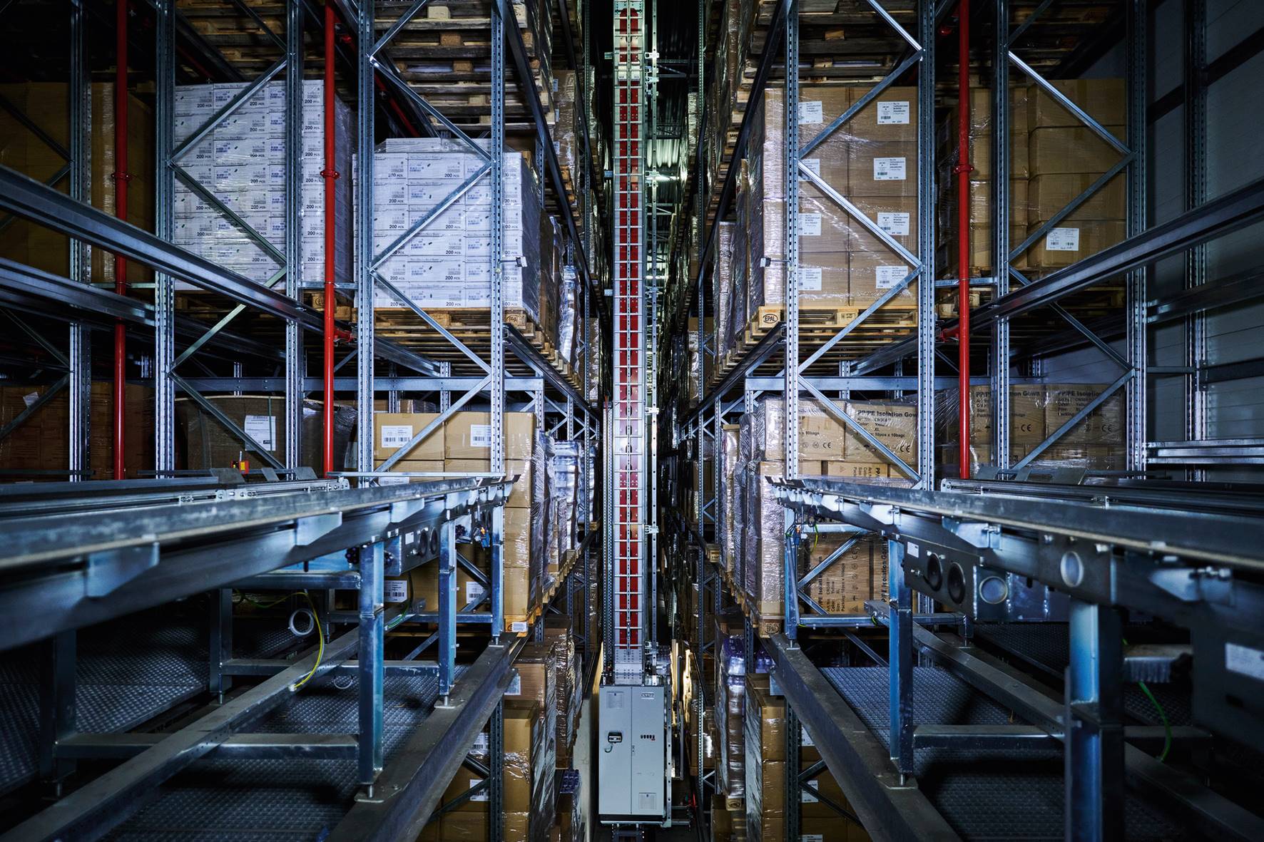 Ein modernes Lager mit hohen Regalen, in denen sich Kisten stapeln, und einem automatischen Regalbediengerät, das sich zwischen den Gängen bewegt. Die Umgebung ist industriell und organisiert, mit Metallstrukturen und sichtbaren Produktetiketten.