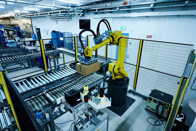 A yellow robotic arm places a cardboard box on a conveyor belt in a modern pharmaceutical distribution warehouse with automated machinery.
