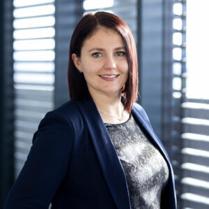 Eine Frau mit glattem, dunklem Haar, die einen marineblauen Blazer und eine gemusterte Bluse trägt, steht lächelnd neben den Jalousien – durch die das Licht hereinströmt – und ist Expertin für AMR-Integration.