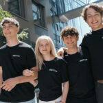 Vier junge Erwachsene stehen im Freien, lächeln und tragen passende schwarze "KNAPP Crew"-T-Shirts.