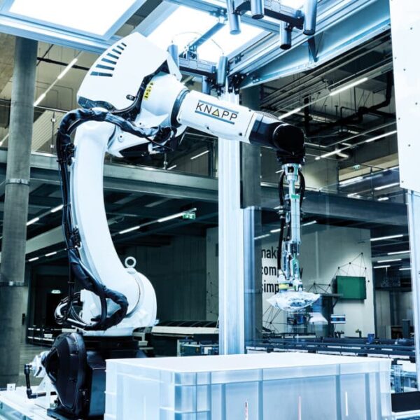 BN Lichtbilder Ein Pick it Easy-Roboter bewegt einen Gegenstand über einen durchsichtigen Behälter in einer modernen Fabrik, umgeben von Glaswänden und Industrieanlagen.