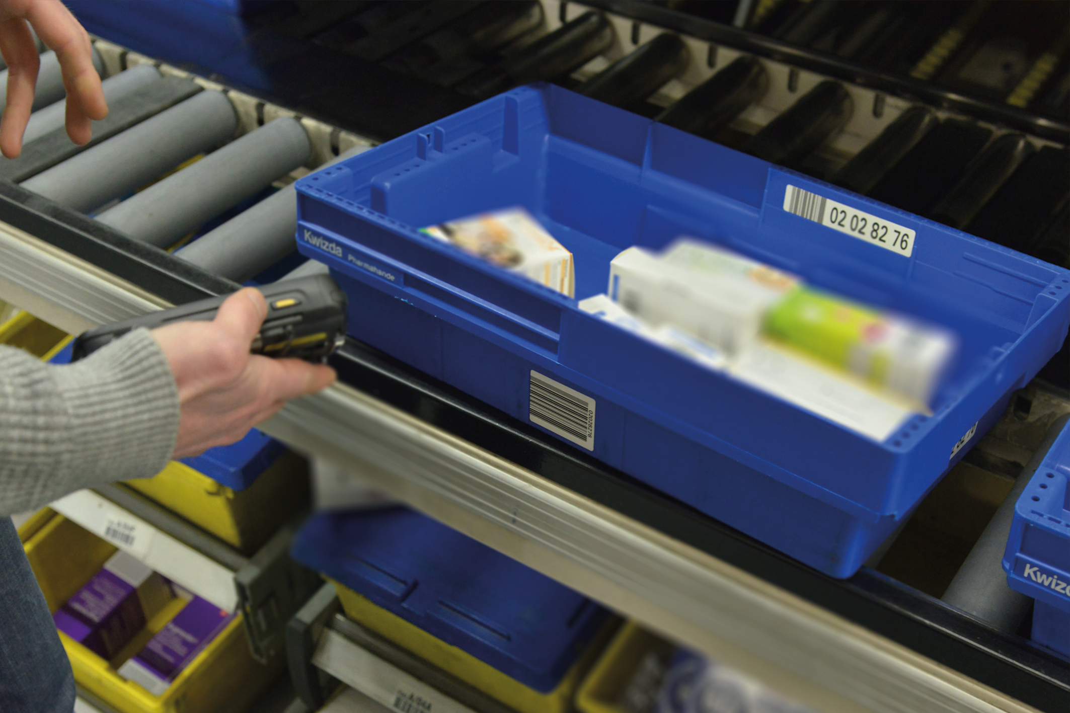 Una persona escanea un contenedor azul lleno de diversos artículos utilizando un lector de códigos de barras manual en un almacén o instalación de clasificación.