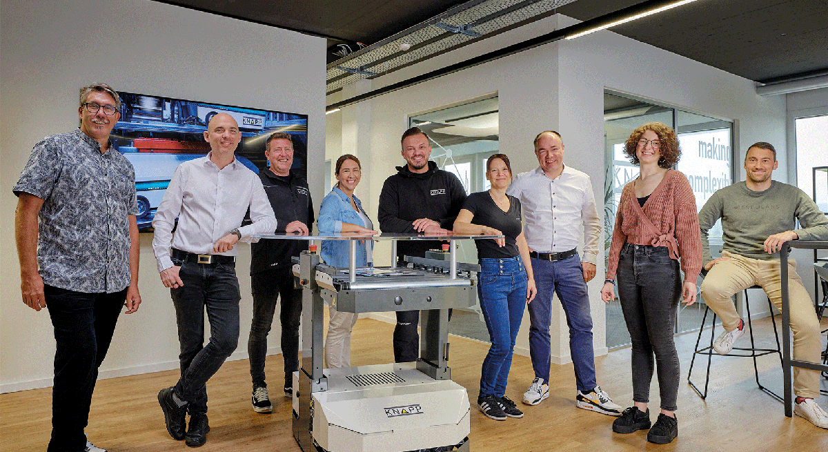 Eine Gruppe von neun Personen, die lächelnd in einem modernen Büroraum von KNAPP Schweiz zusammen posieren, steht um ein kleines Robotergerät auf Rädern. Einige stehen, andere sitzen auf hohen Hockern.