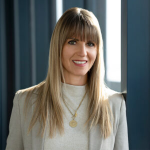 Eine lächelnde Frau mit glattem blondem Haar steht vor blauen Tafeln, trägt einen hellen Blazer und eine Halskette und repräsentiert KNAPP Industry Solutions.