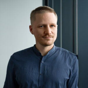 Ein Mann mit kurzem blondem Haar und gestutztem Bart, der einen blauen Button-up trägt, steht an einem Fenster und repräsentiert KNAPP Industry Solutions mit einem leichten Lächeln.