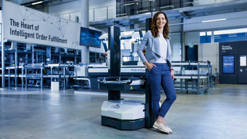 Eine Frau lächelt neben einem autonomen Roboter in einem modernen Lagerhaus, das KNAPP Industry Solutions und "The Heart of Intelligent Order Fulfillment" vorstellt.