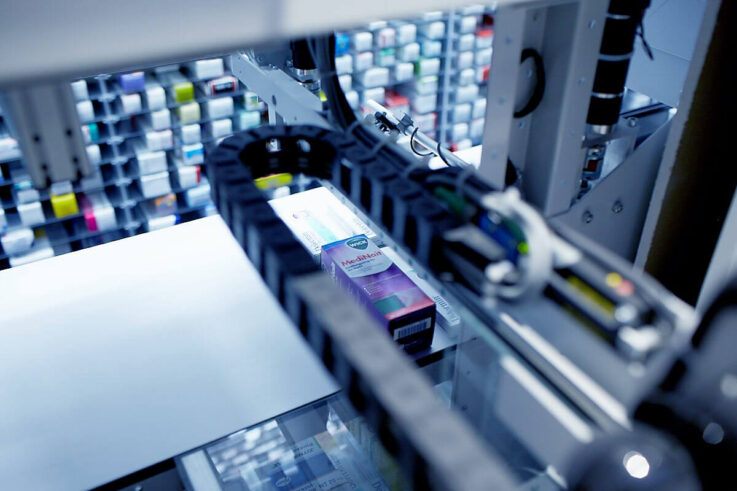 Kommisionierautomat-Technik_A2000-A3000_Greifer_Greifarm_Tablartechnik_Puffer-737x491 Un brazo robótico manipula una caja de medicamentos en una farmacia, haciendo gala de logística sostenible entre estanterías repletas de diversos envases de medicamentos.