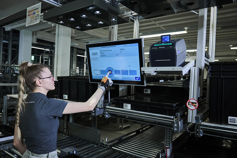 Picking at the ergonomic work stations at B&R A young woman is touching the screen above the picking work station which is connected to a conveyor system.