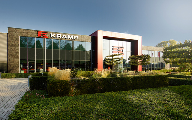 Modern office building with large glass windows and a bold red entrance frame, surrounded by manicured green hedges and trees. The sign "KRAMP" is displayed on the front of the building.