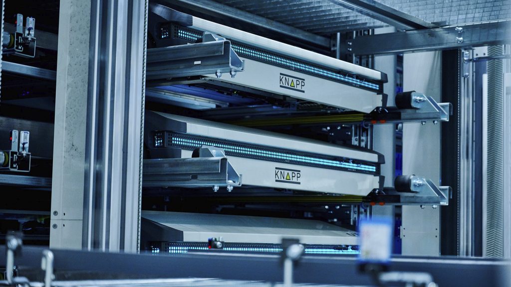 Soluciones de almacenamiento para la industria del plástico Tres sistemas Shuttle con el logotipo de KNAPP e iluminación azul se encuentran en un sistema de almacenamiento.