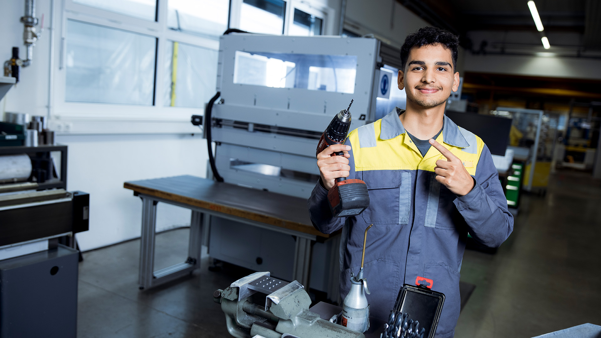 An apprentice with a tool in his hand smiles into the camera with commitment.
