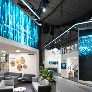 Moderner Messestand mit digitalen Bildschirmen, weißen Möbeln und blauer Beleuchtung, der Automatisierungslösungen für die gesamte Wertschöpfungskette präsentiert.