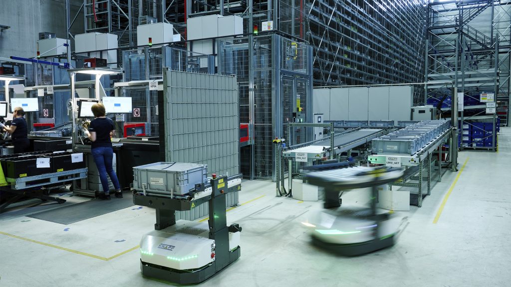 Dos robots móviles autónomos se dirigen hacia un transportador, un sistema de almacenamiento y puestos de trabajo.
