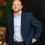 Ein Mann in blauem Blazer und Jeans steht in einem Innenraum und lächelt mit einer Hand in der Tasche vor einer dunklen Wand.