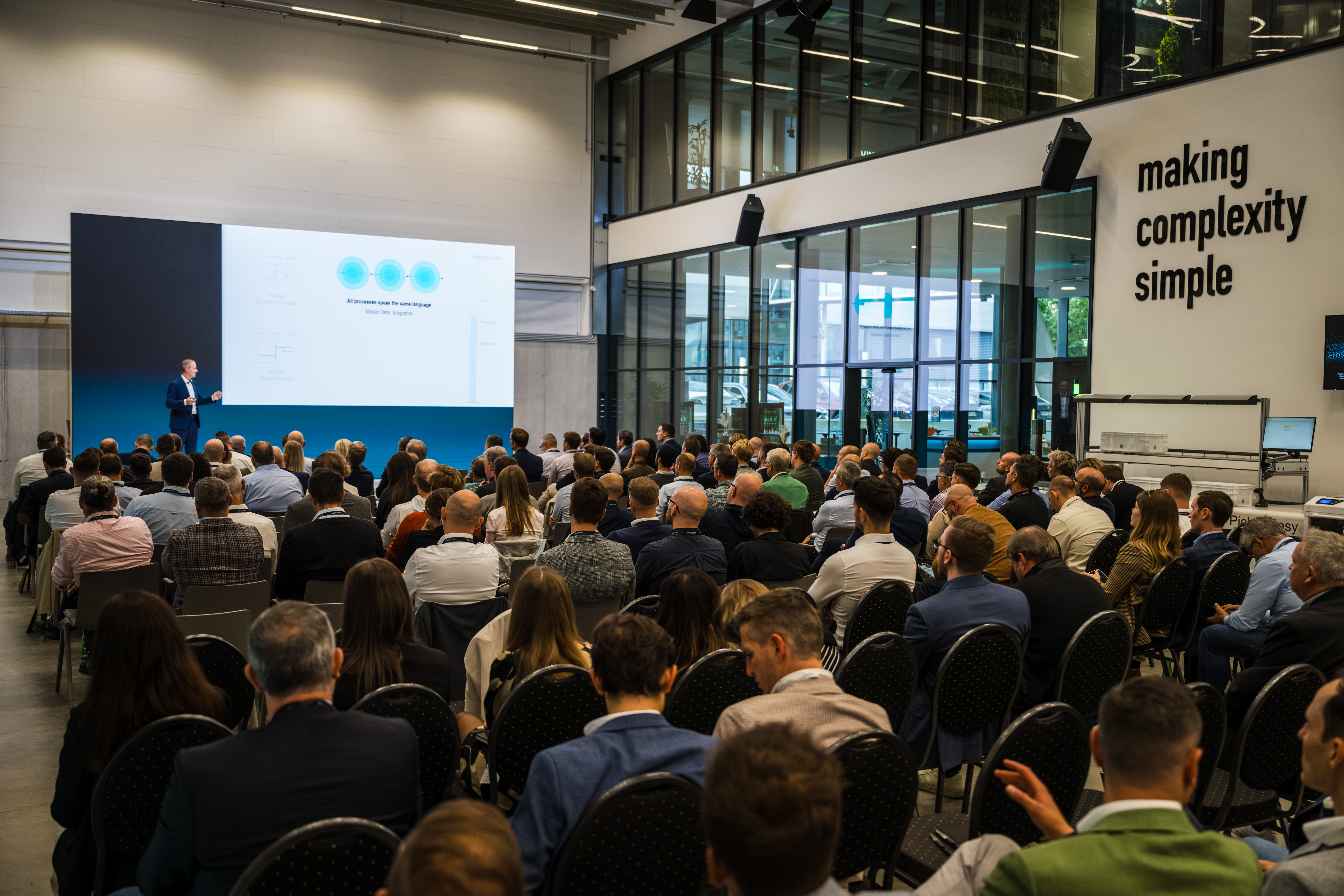 events-knapp-ag Ein großes Publikum verfolgt einen Vortrag mit Folien in einem modernen Konferenzraum, während die Schlagzeile "Komplexität einfach machen" in den Nachrichten erscheint.
