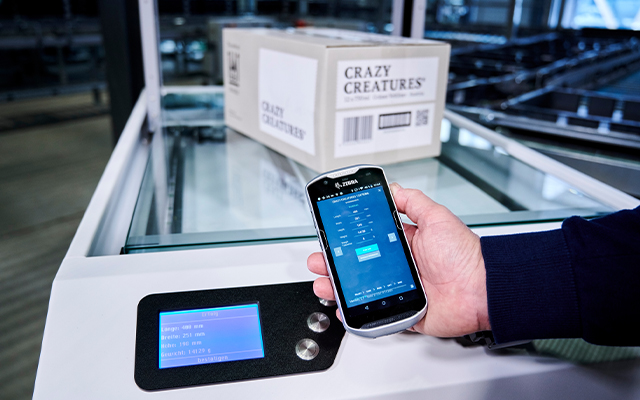 multiscan_automatische_stammdaten_erfassung Une personne tient un lecteur de codes-barres avec une interface numérique près d'un panneau de commande, avec une boîte étiquetée "Crazy Creatures" et un code-barres en arrière-plan sur un système de convoyage dans un entrepôt.