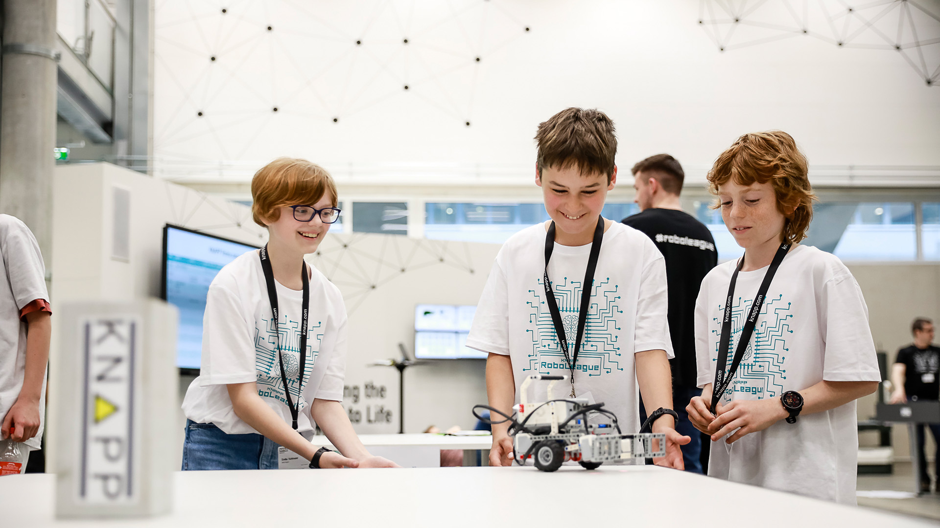 Three young pupils enthusiastically watch their robot move at a RoboLeague event.