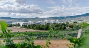 Inmitten grüner Felder und Bäume steht eine große weiße Industrieanlage, im Hintergrund sind Hügel und ein teilweise bewölkter Himmel zu sehen.