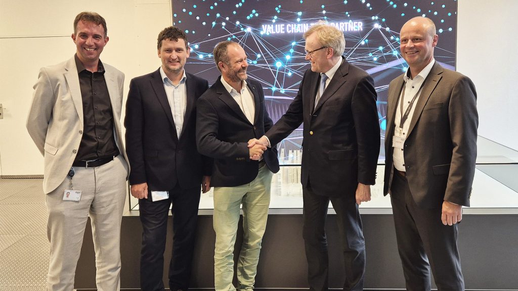 Cinq hommes en tenue de travail se tiennent ensemble, deux au centre se serrant la main et souriant, devant un écran affichant un graphique de réseau numérique et les mots "VALUE CHAIN PARTNER".